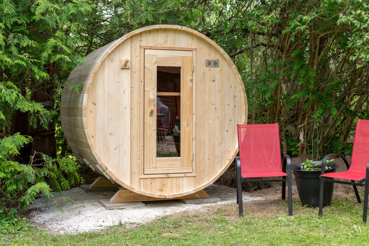 CT Harmony barrel sauna front view set on a gravel pad against a cedar hedge with patio seating