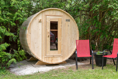 CT Harmony barrel sauna front view set on a gravel pad against a cedar hedge with patio seating
