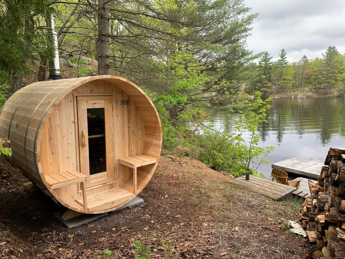 CT Serenity barrel sauna installed lakeside with wood-burning chimney and firewood stack