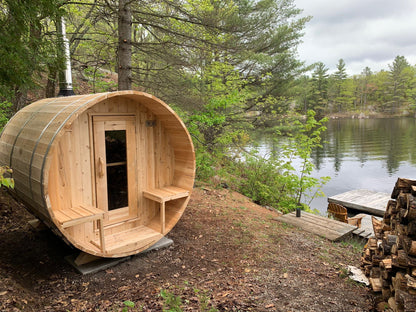 CT Serenity barrel sauna installed lakeside with wood-burning chimney and firewood stack